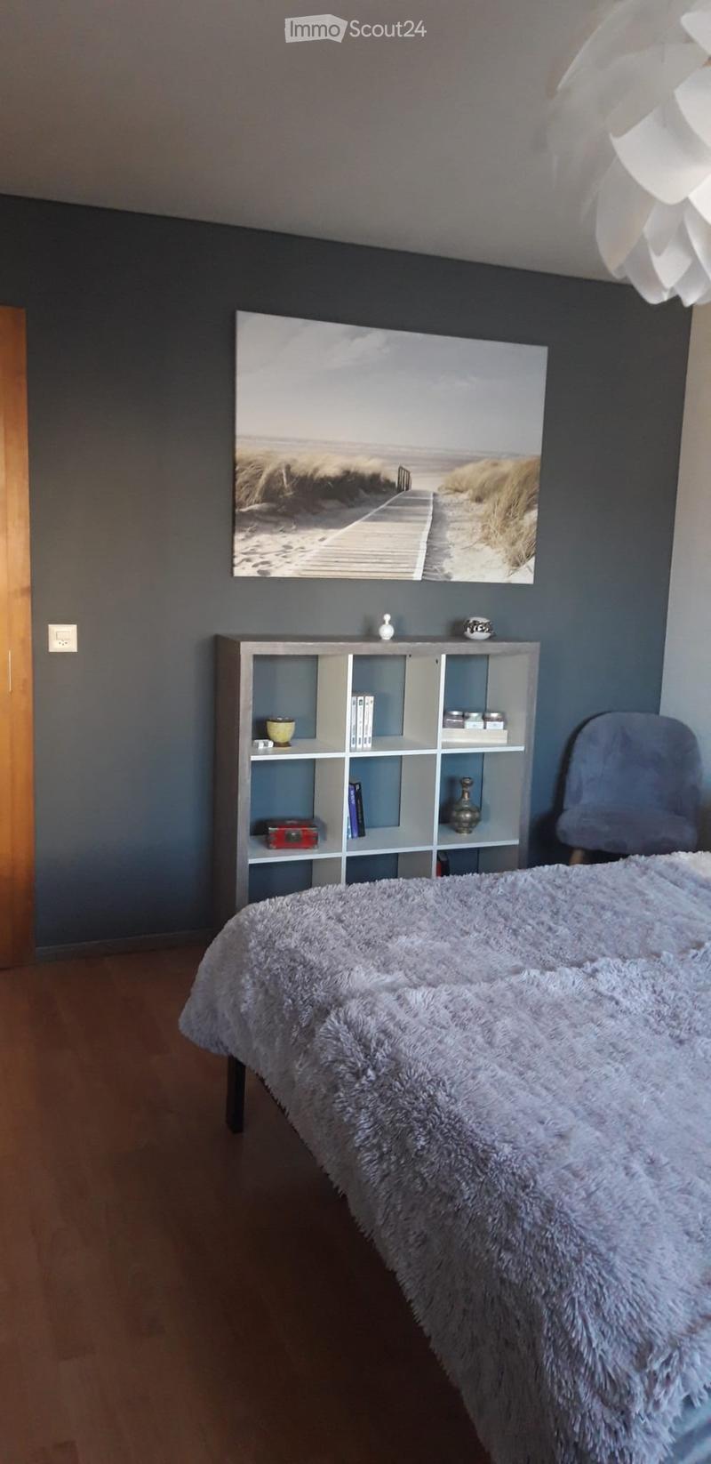 Modern bedroom with a large bed covered in a fluffy white blanket, a grey armchair, a white shelving unit with books and decorative items, and a large picture frame on the wall.