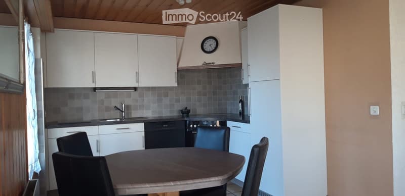 Modern kitchen with white cabinets, a round wooden dining table with four black chairs, a sink with a faucet, an oven, and a dishwasher. A clock is mounted on the wall above the stove.