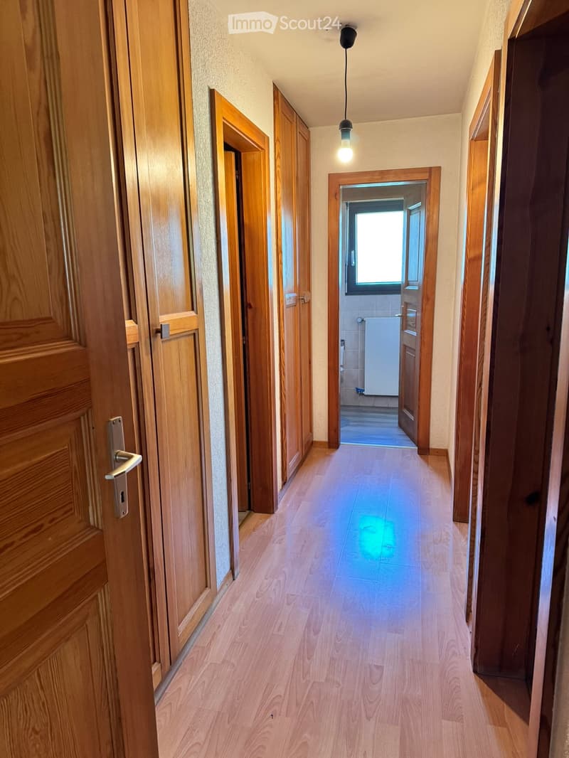 A well-lit hallway with wooden flooring and light-colored walls, featuring several wooden doors and a visible bathroom at the end.