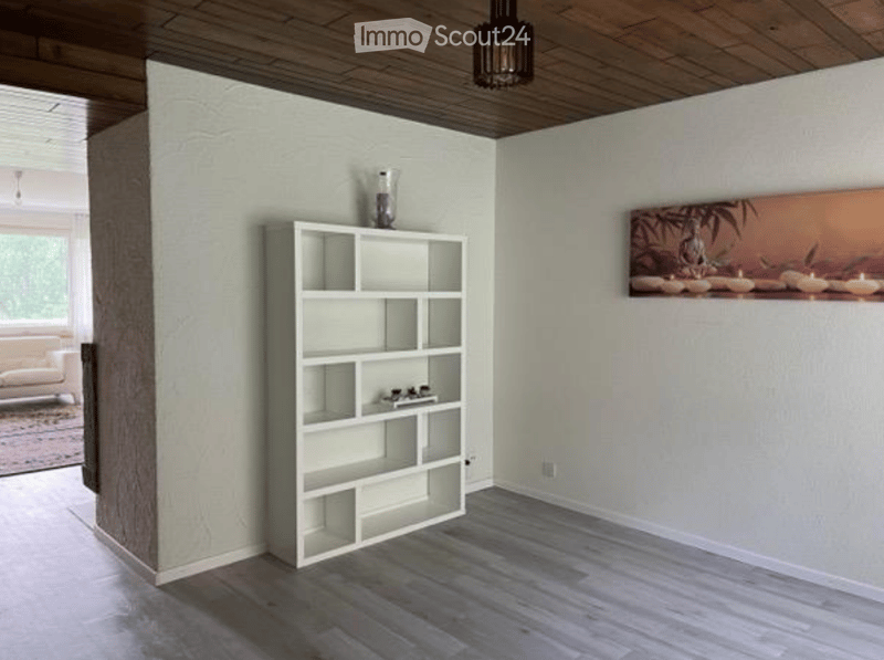 Modern, minimalist room with light grey walls, dark wood flooring, and a white open shelving unit. A large painting hangs on one wall, and a sofa is visible in the adjacent space.