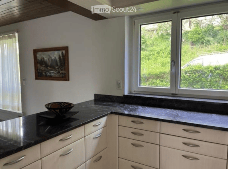 Modern kitchen with light wood cabinets, black granite countertops, and a decorative bowl on the counter. Large windows provide natural light and a view of greenery outside.