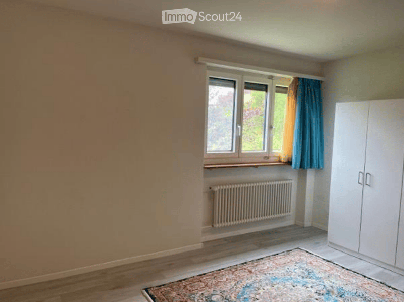 Empty room with wooden flooring, white walls, large window with blue curtains, radiator, and a white wardrobe.