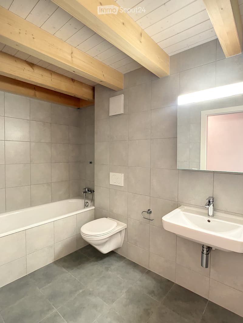 Modern bathroom with a bathtub, toilet, sink, and mirror. Exposed wooden beams on the ceiling and grey tiled walls.