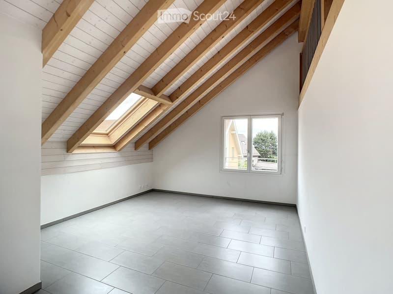 Empty room with white walls, grey tiled floor, exposed wooden beams, skylight windows, and a large window with a view of the outside.