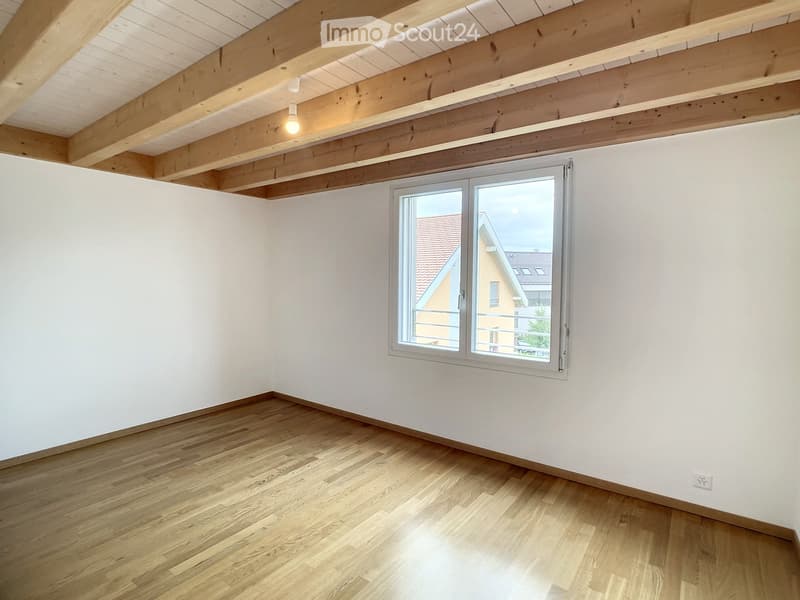 Empty room with wooden floor, white walls, and exposed wooden ceiling beams. A large window offers a view of the outside.