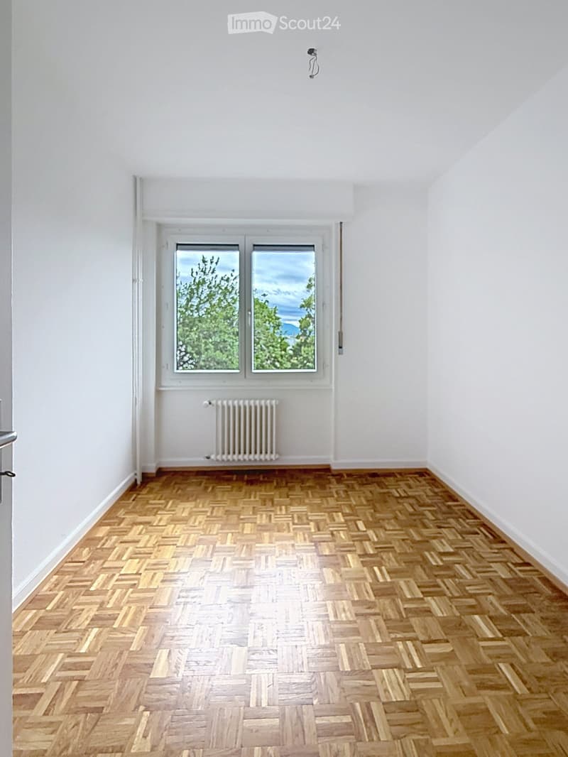 Empty room with wooden herringbone flooring, white walls, and large windows showing greenery outside. A radiator is visible below the windows.