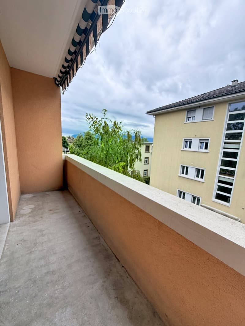 Balcony with concrete floor, beige walls, and a view of neighboring buildings and greenery.