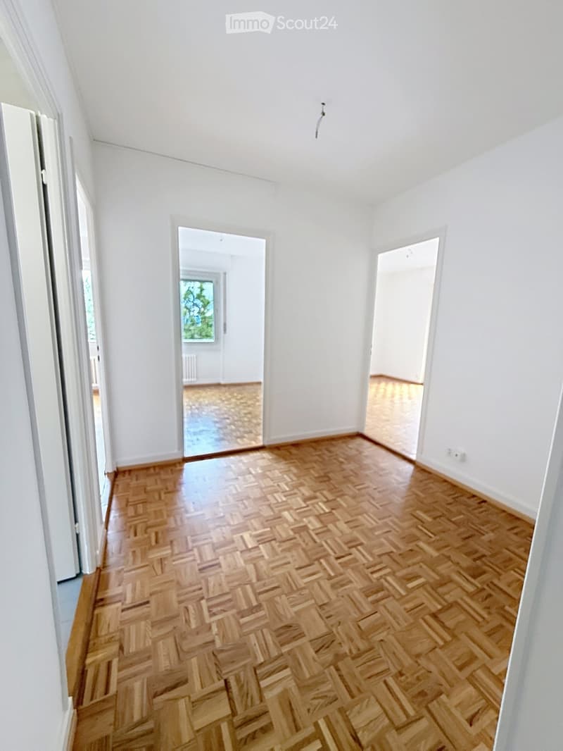 Empty room with white walls and herringbone wooden flooring, featuring two open doorways leading to other spaces.