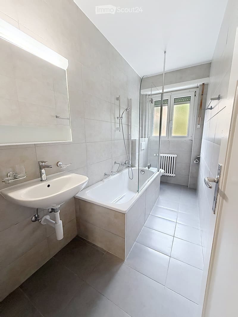 Modern bathroom with a white sink, bathtub with glass shower enclosure, mirror, and radiator.