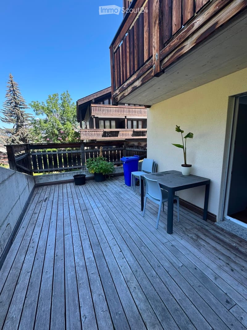 Wooden balcony with a table, chairs, potted plants, and a blue trash bin.