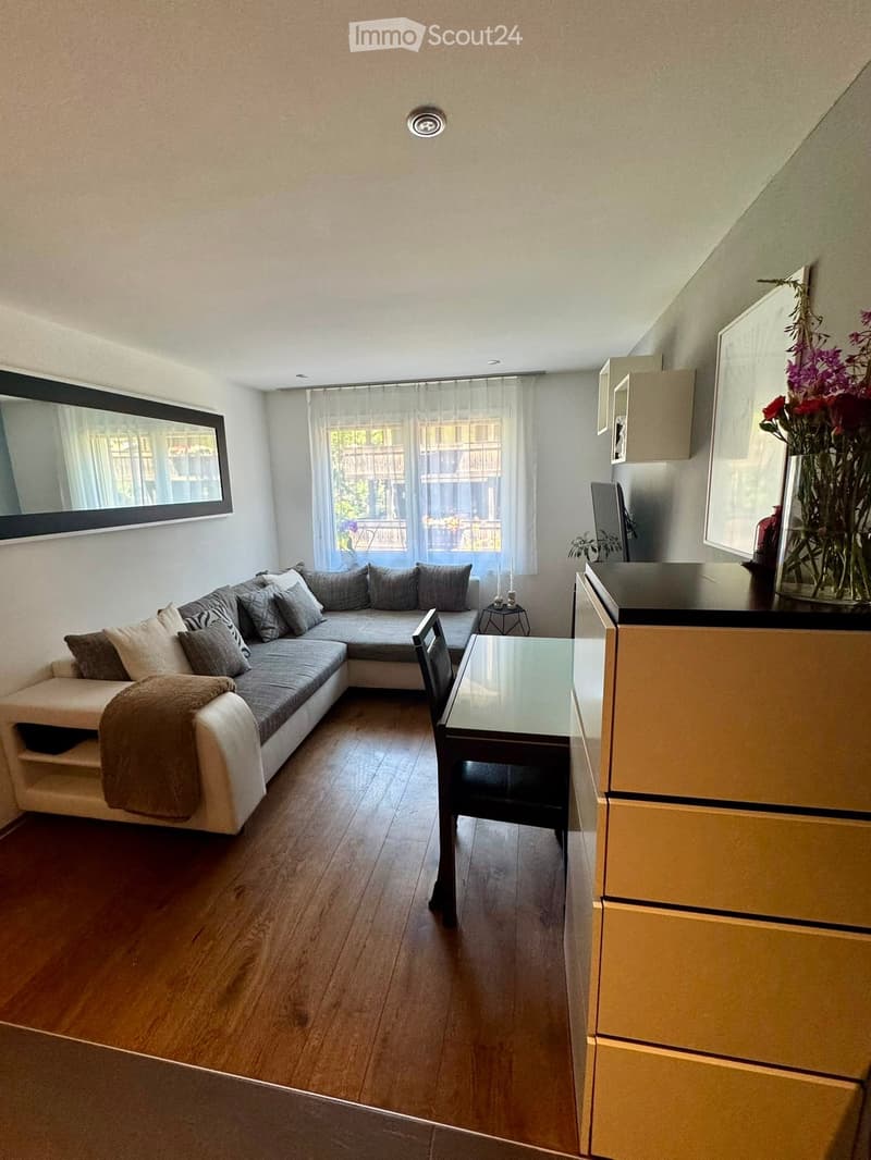 A modern living room with a sectional sofa, a glass dining table, a wooden chair, a vase with flowers, a mirror, and a candle holder. The room features wooden flooring and large windows with sheer curtains.