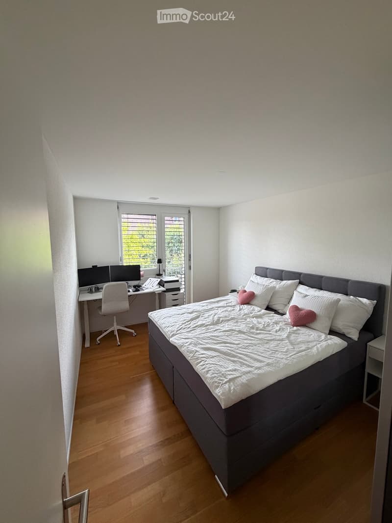 Modern bedroom with a large bed, white bedding, two pink heart-shaped pillows, a desk with a computer and chair, and a window with blinds.