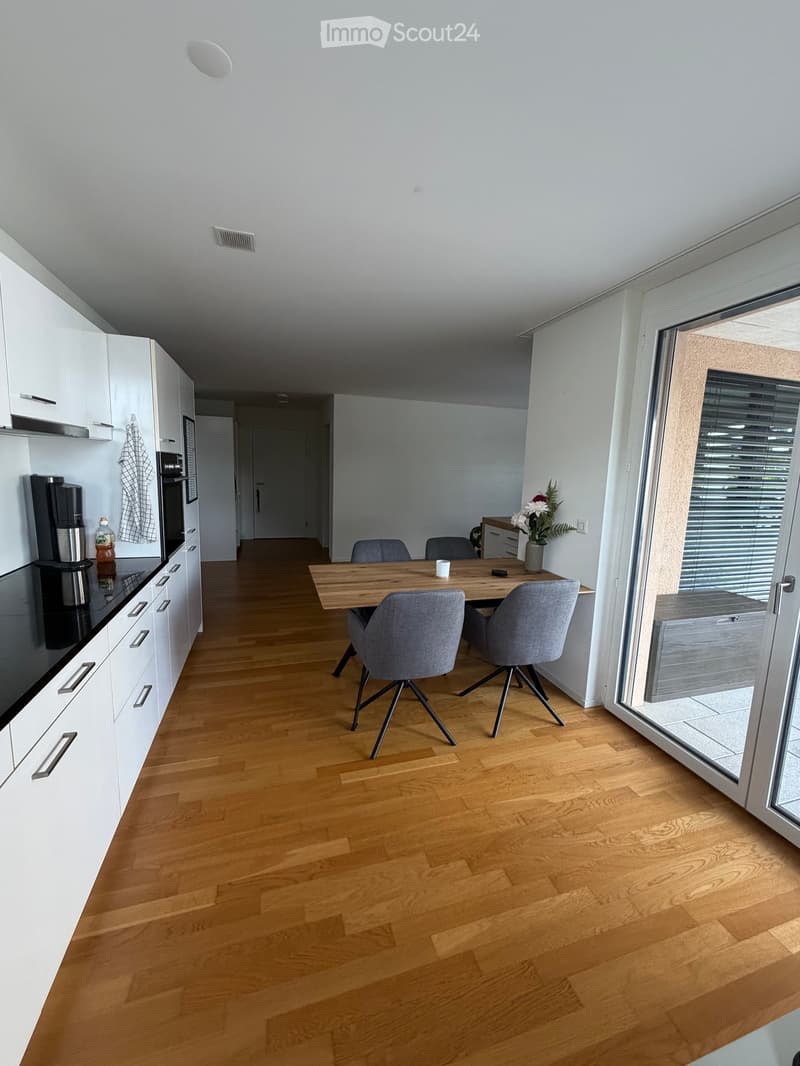 Open-plan kitchen and dining area with white cabinets, a coffee maker, an oven, a wooden dining table, four grey chairs, and a vase with flowers on the table.