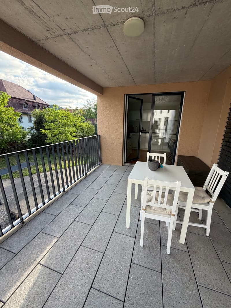 Balcony with tiled floor, white table and chairs, overlooking a green area with trees and houses.
