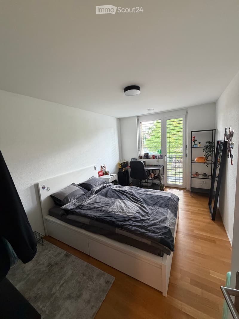 Modern bedroom with a large bed, desk, chair, mirror, and shelf. The room features wooden flooring and large windows with blinds.