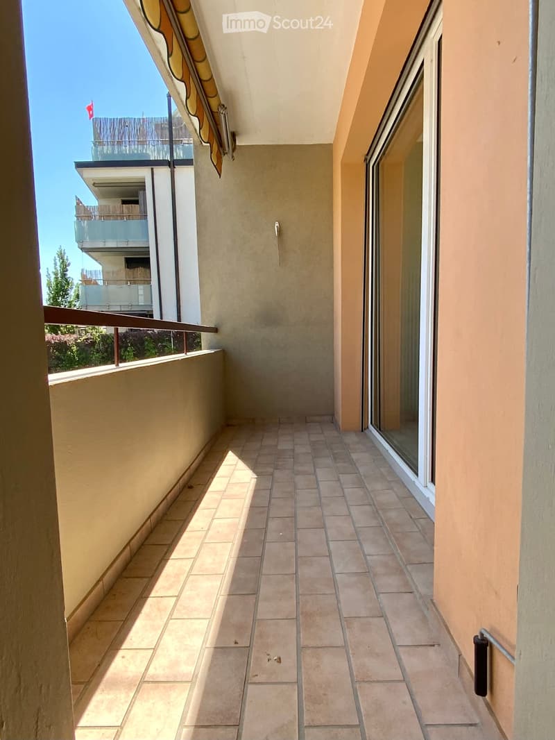 Balcony with tiled floor, glass railing, and view of neighboring building and greenery.