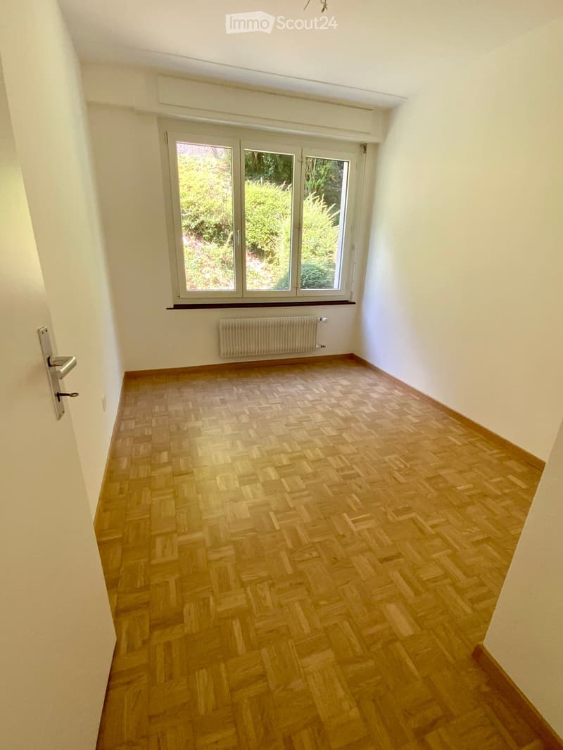 Empty room with wooden herringbone flooring, white walls, large window with view of greenery, radiator, and door handle visible.