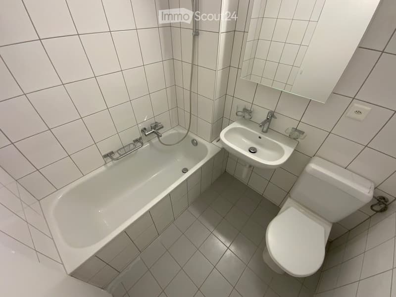Clean bathroom with white tiled walls and floor, featuring a bathtub, toilet, sink, and mirror.