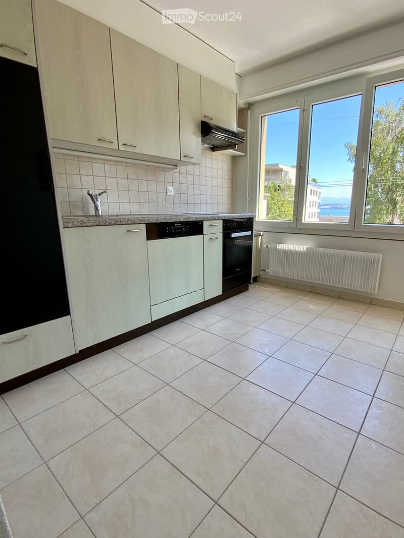 Modern kitchen with light-colored cabinets, tiled floor, and large windows offering a view of the sea and surrounding buildings.