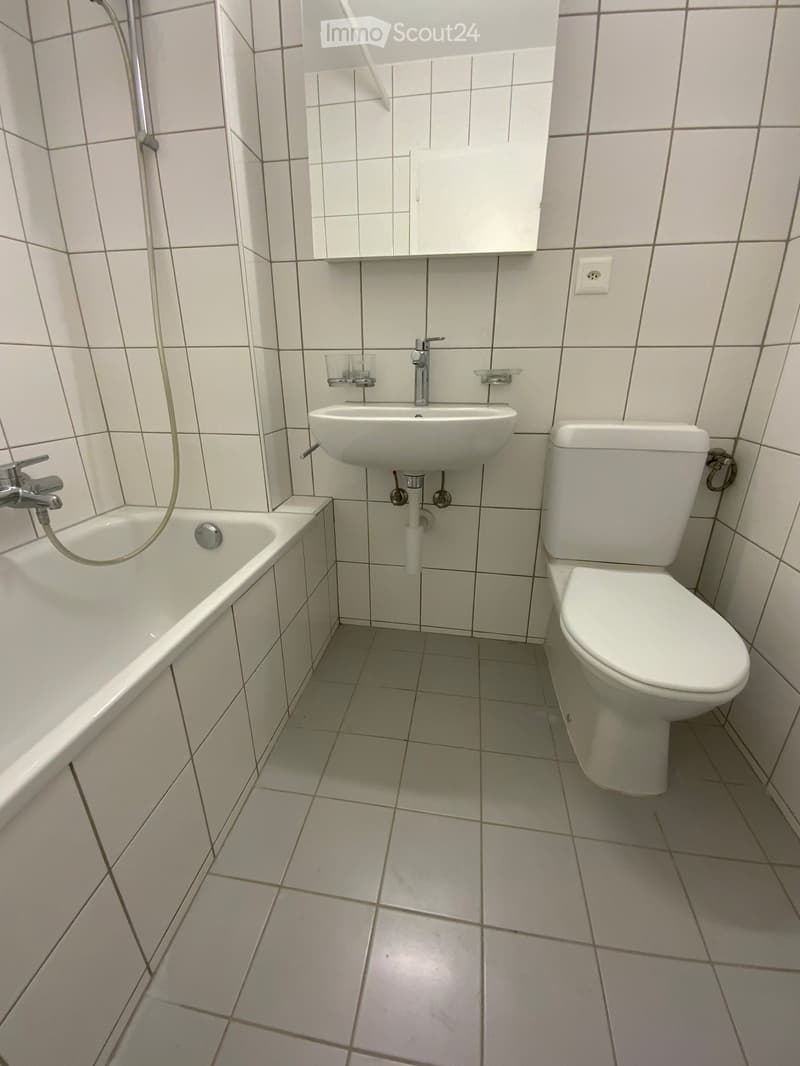 Clean, tiled bathroom with a bathtub, toilet, sink, and mirror. The walls and floor are covered in white tiles, and the fixtures are modern and white.