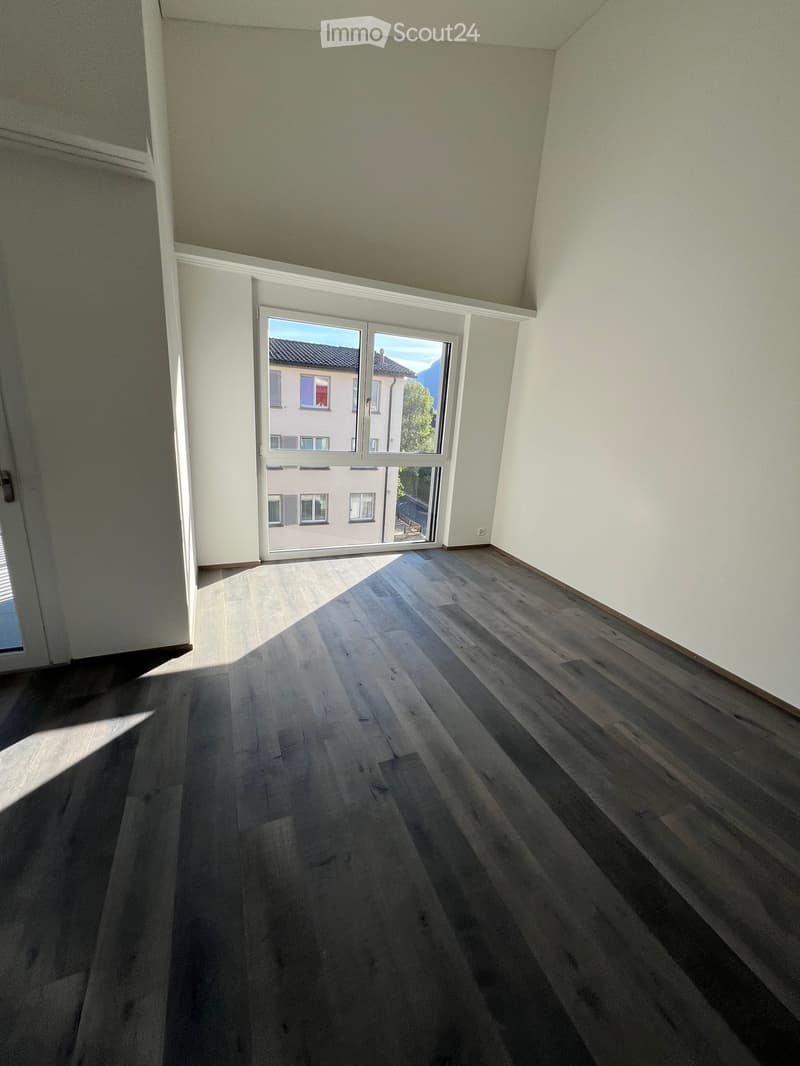Empty room with wooden flooring and large windows, allowing natural light to enter. The walls are painted in a light color, and the ceiling has a slightly sloped design.