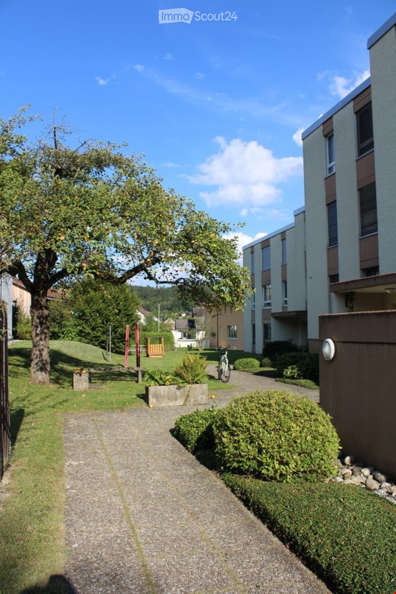 Well-maintained garden area with a tree, pathway, and modern apartment building in the background.