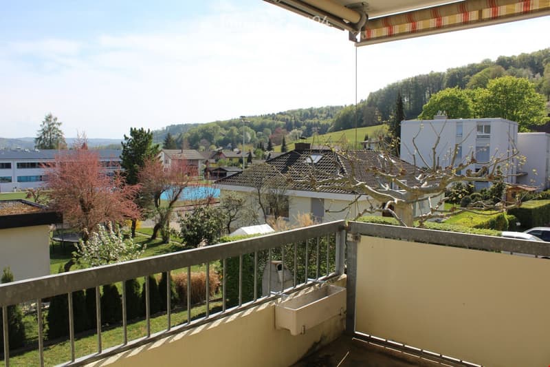 Balcony with metal railing overlooking a residential area with houses, trees, and a swimming pool in the distance.