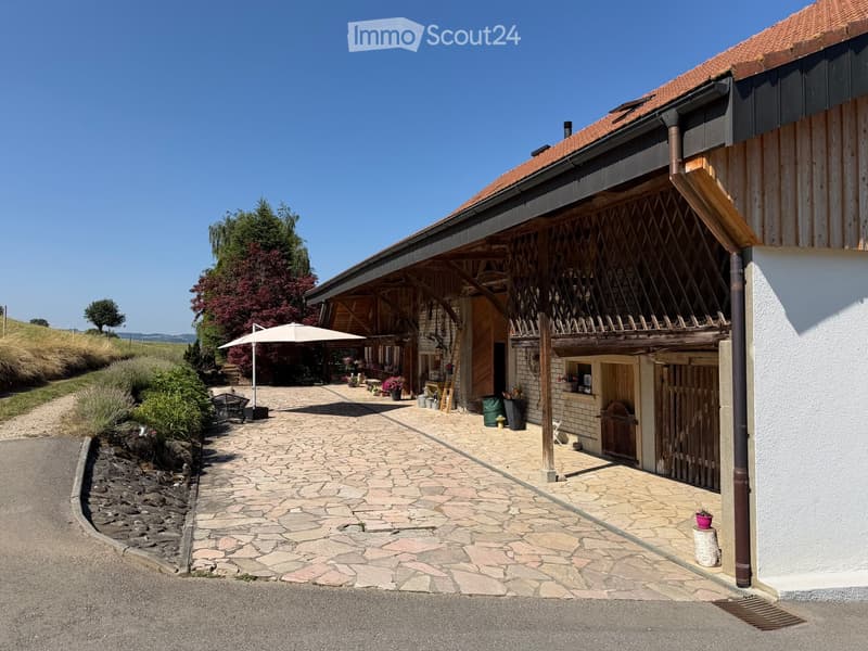 A rustic, two-story wooden building with a red-tiled roof, stone-paved courtyard, and outdoor seating area with an umbrella.