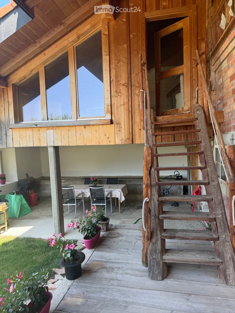 Wooden deck with a table, chairs, flower pots, and a staircase leading up to a wooden balcony with large windows.