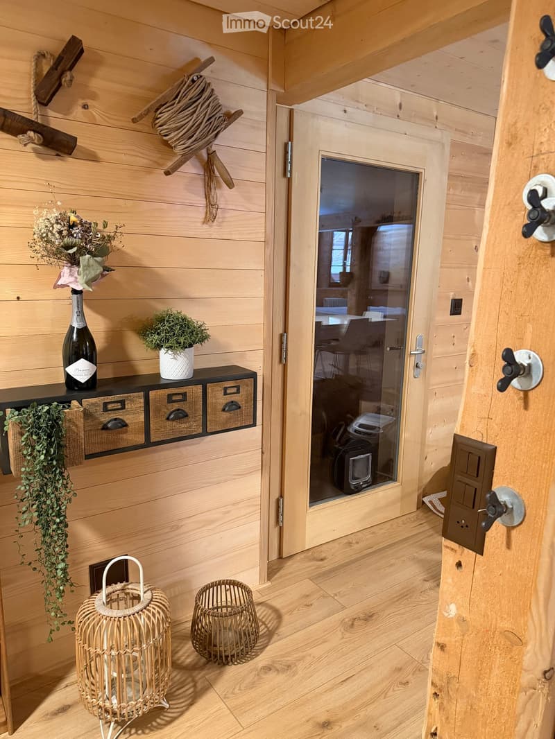 Cozy wooden room with a shelf holding a bottle, flower pot, and drawers, along with wicker baskets and a cat litter box visible through an open door.