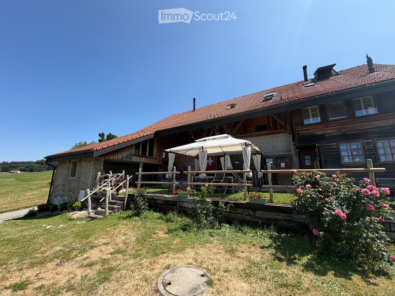 A two-story wooden house with a tiled roof, surrounded by a grassy field and a wooden deck with outdoor furniture and an umbrella.