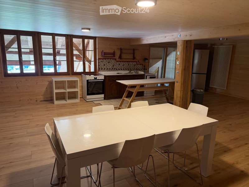 Open-plan room with wooden flooring, white dining table with chairs, small white shelf, oven, refrigerator, and large windows.