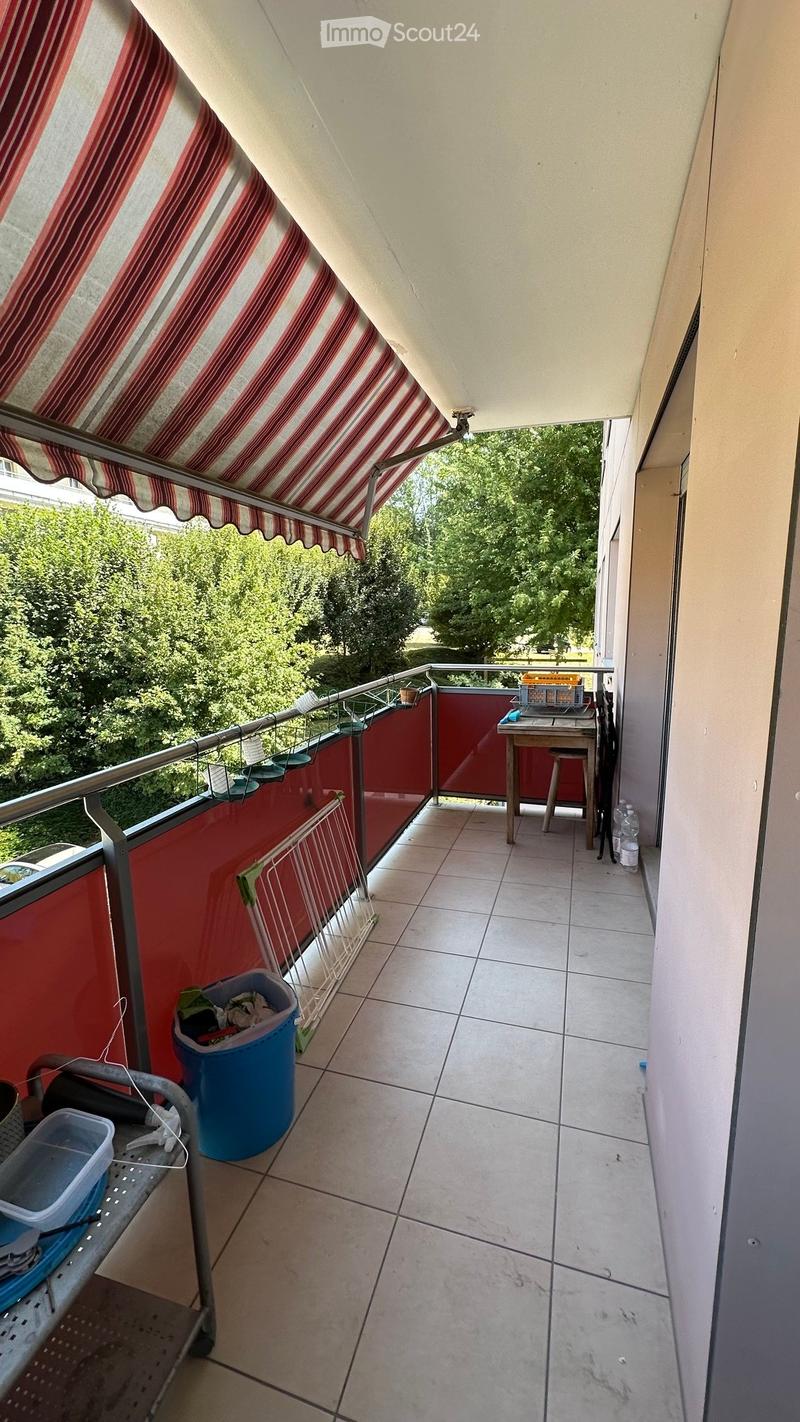 Balcony with tiled floor, metal railing, and striped awning. Includes a small table, stool, laundry rack, and trash bin. Offers a view of greenery outside.