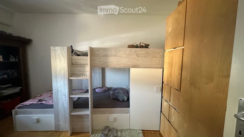 Cozy bedroom with a lofted twin-sized bed, built-in wardrobe, and shelving unit. The lower bunk has a mattress and pillows, and the room features wooden flooring and a small rug.