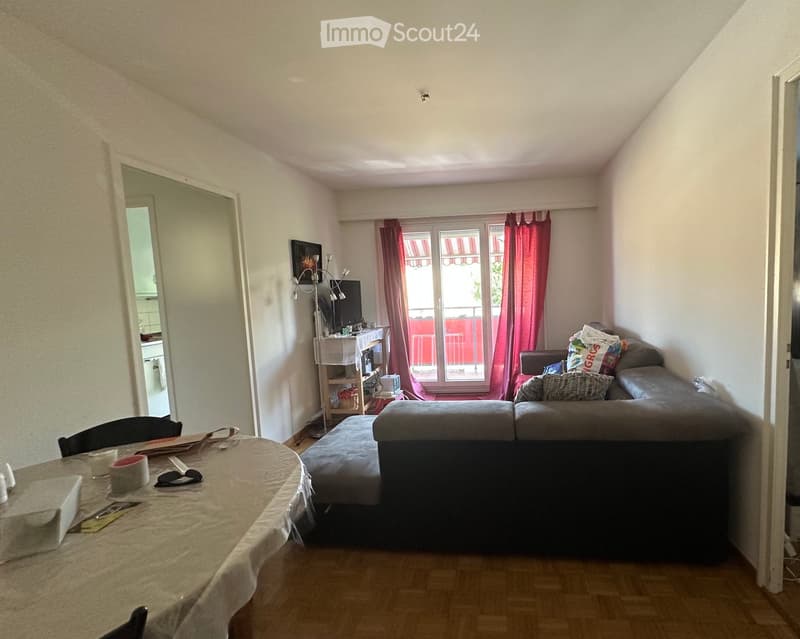 A furnished living room with a sofa, dining table, chairs, television, shelf, and mirror. The room has wooden flooring and large windows with red curtains, leading to a balcony.