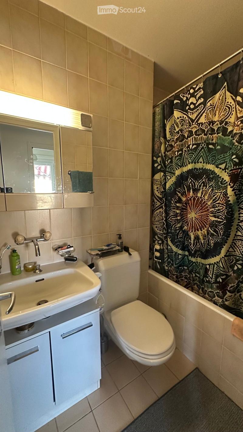 Bathroom with beige tiled walls, a white sink with a mirror cabinet, a toilet, and a shower area with a colorful curtain.