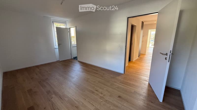 Empty room with light wooden flooring and white walls, featuring two open doors leading to other rooms and a window with a view of greenery.