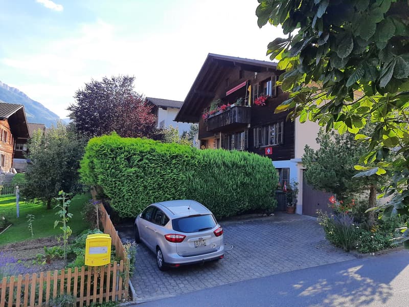 2 story house, wooden exterior, balcony with flowers, car parked in front, garden with hedges and plants