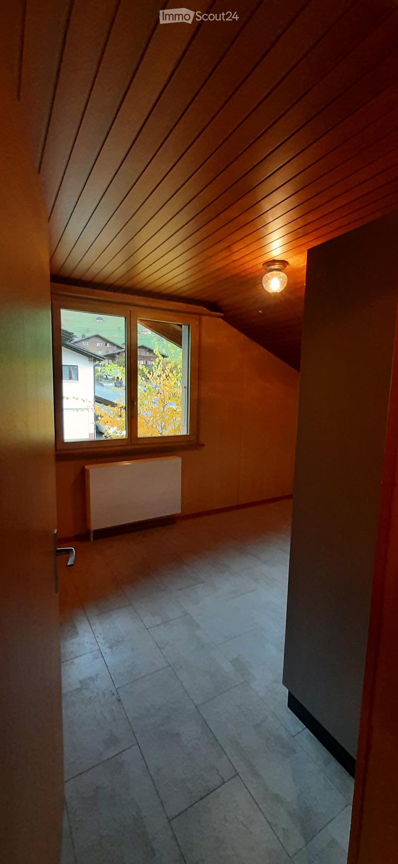 Empty room with wooden walls, slanted wooden ceiling, tiled floor, radiator, and large window showing outdoor view.
