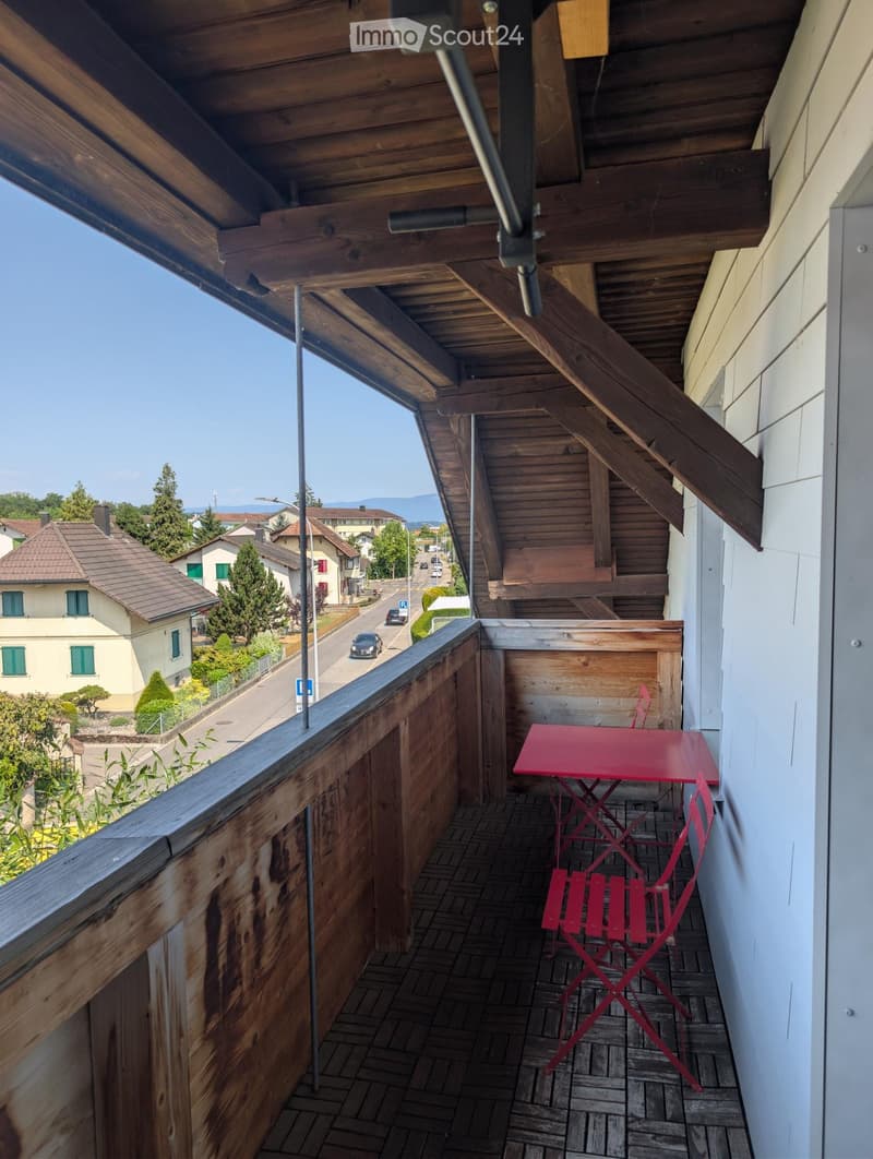 Balcony with wooden railing, tiled floor, red table and chairs, overlooking a street with houses and trees.