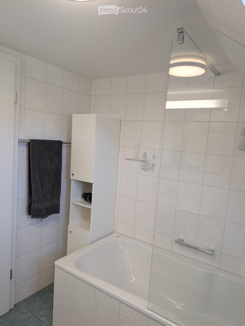 Modern bathroom with white tiled walls, a bathtub, a white cabinet, a towel on a rack, and a shelf with decorative items.
