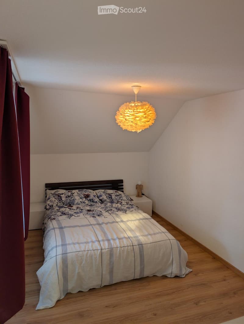 Cozy bedroom with wooden flooring, slanted ceiling, and a modern hanging light fixture. Features a double bed with a plaid duvet and matching pillows, a nightstand with a small lamp, and a dark wooden headboard.