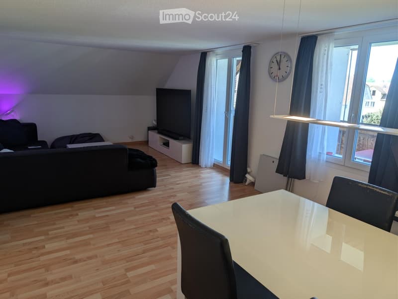 Modern living room with wooden flooring, large windows, a sofa, a dining table with chairs, and a television on a stand.