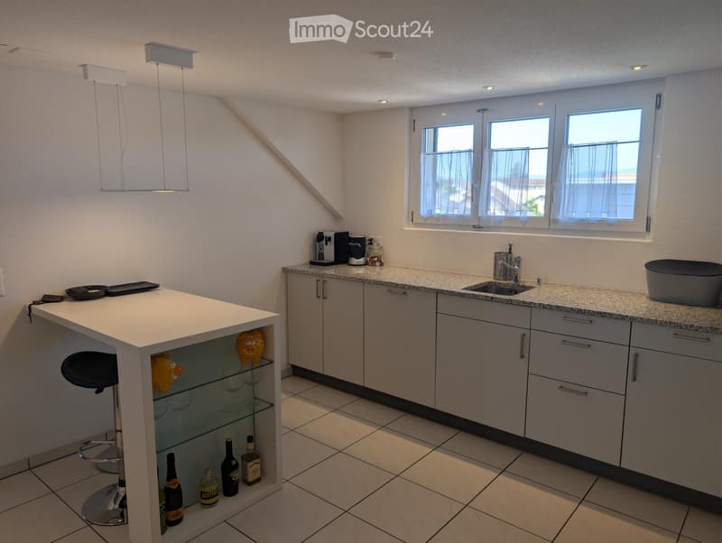 Modern kitchen with grey cabinets, granite countertops, and a white island with glass shelves holding wine glasses and bottles. A bar stool is positioned at the island, and a coffee maker sits on the counter. The room features pendant lighting and large w