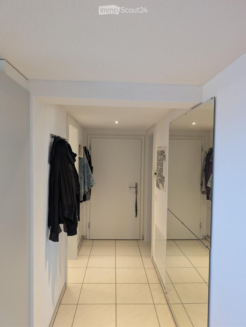 A clean, well-lit hallway with tiled flooring, white walls, and several doors leading to different rooms. A coat rack with jackets is visible on the left side.