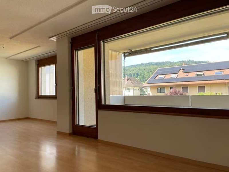 Empty room with wooden flooring, large windows, and a sliding glass door leading to a balcony. The walls are painted white, and the room has a modern, minimalist design.