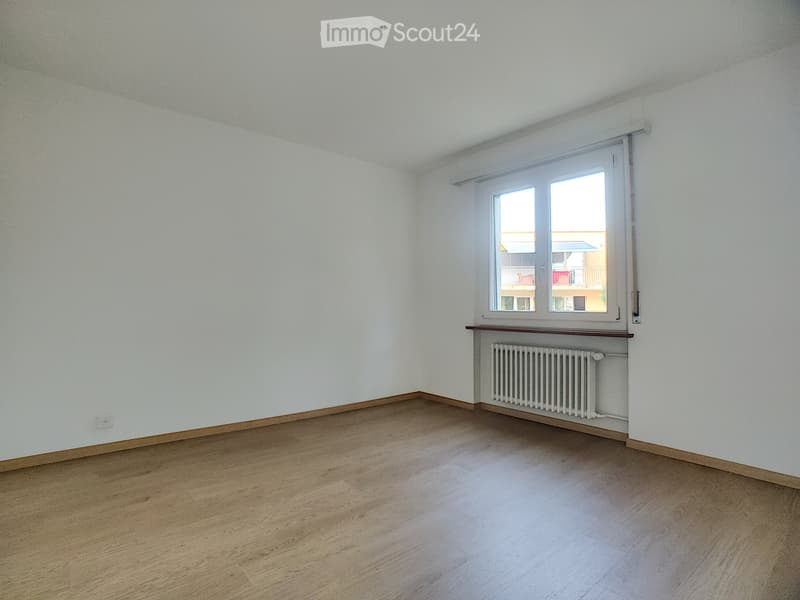 Empty room with light wooden flooring, white walls, and a window with a radiator below it.
