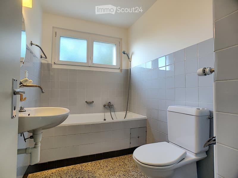 Modern bathroom with a white sink, bathtub, toilet, and showerhead. The walls are tiled in light gray, and there is a window above the bathtub.