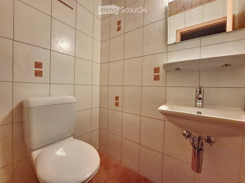 A small bathroom with white tiled walls and a wooden floor. It features a toilet, a sink with a faucet, and a mirror above the sink.