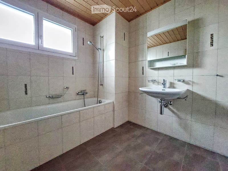 Modern bathroom with a bathtub, sink, mirror, and showerhead. Light-colored tiles and wooden ceiling.
